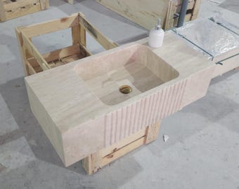 May include: A rectangular beige stone sink featuring a smooth basin and a front panel with vertical fluted detailing. The sink includes a central gold-toned drain cover and a white soap dispenser placed on the flat top surface of the stone unit.