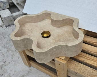 May include: A beige travertine stone sink featuring a distinctively wavy, organic-shaped rim and a smooth, polished surface. The basin includes a centred, circular gold-toned metal drain cover, showcasing the natural porous texture of the stone.