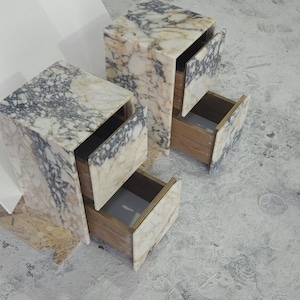 May include: Two marble-patterned bedside tables with open drawers. The tables have a rectangular shape with a mix of white, beige, and gray veining. The drawers are made of wood. The tables are on a gray surface.