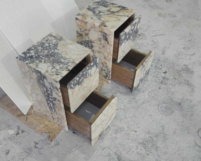 May include: Two marble-patterned bedside tables with open drawers. The tables have a rectangular shape with a mix of white, beige, and gray veining. The drawers are made of wood. The tables are on a gray surface.