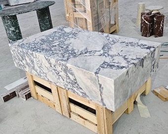 May include: A large rectangular marble block with a white and gray veined pattern. The block is supported by a wooden pallet. Other marble pieces and a green marble table are visible in the background.