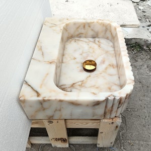 May include: A rectangular marble sink with a cream and brown veined pattern. The sink has a rounded interior and a decorative fluted edge. A gold-colored drain cover is visible in the center of the basin. The sink is resting on a wooden pallet.