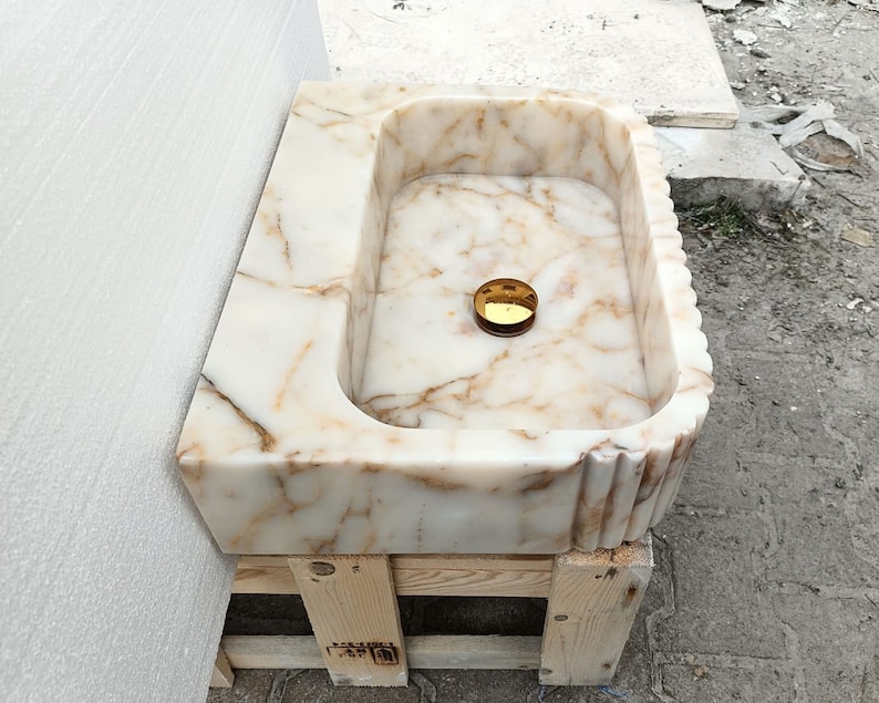 May include: A rectangular marble sink with a cream and brown veined pattern. The sink has a rounded interior and a decorative fluted edge. A gold-colored drain cover is visible in the center of the basin. The sink is resting on a wooden pallet.