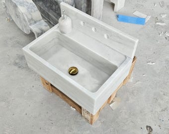 May include: A rectangular white marble sink featuring a high back splash with three pre-drilled faucet holes. The basin includes a polished gold-toned drain cover and a small white ceramic soap dispenser resting on the top ledge of the sink unit.