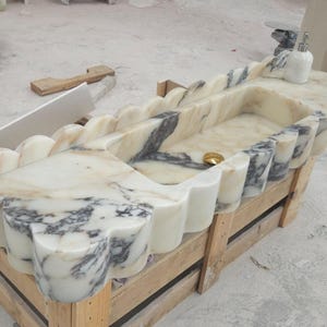 Puede incluir: Un lavabo rectangular de m&aacute;rmol con borde festoneado y vetas blancas y grises. La cubeta incluye un desag&uuml;e dorado y un dispensador de jab&oacute;n blanco se encuentra en la esquina de la superficie de piedra.