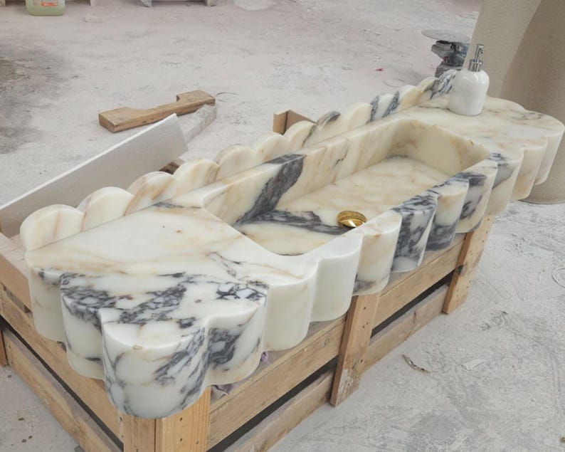 Puede incluir: Un lavabo rectangular de m&aacute;rmol con borde festoneado y vetas blancas y grises. La cubeta incluye un desag&uuml;e dorado y un dispensador de jab&oacute;n blanco se encuentra en la esquina de la superficie de piedra.