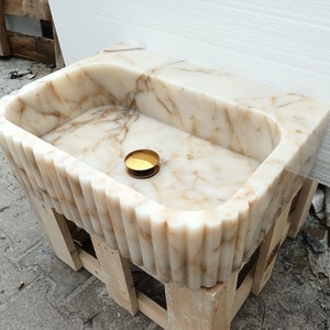 May include: A rectangular marble sink with a fluted front and sides, supported by a wooden frame. The sink features a light beige and brown veined pattern. A small, round, gold-colored object rests inside the basin.