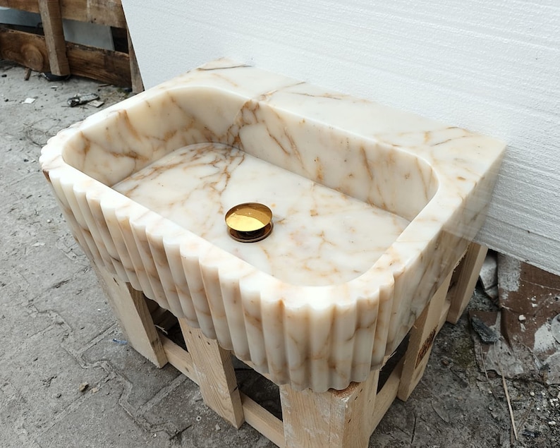 May include: A rectangular marble sink with a fluted front and sides, supported by a wooden frame. The sink features a light beige and brown veined pattern. A small, round, gold-colored object rests inside the basin.