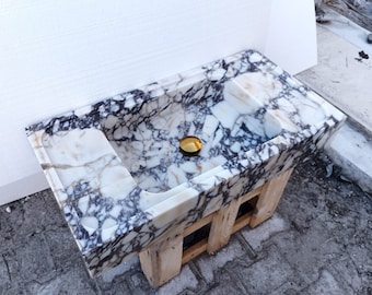 May include: A rectangular marble sink featuring a white base with prominent dark purple and black veining. The basin includes a polished gold-toned circular drain cover and is supported by a light-colored wooden frame structure underneath.