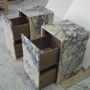 May include: Two marble-patterned nightstands with open drawers. The nightstands have a cream and gray veined pattern. The drawers are made of light-colored wood. The nightstands are rectangular and have a modern design.