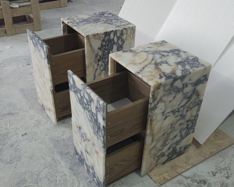 May include: Two marble-patterned nightstands with open drawers. The nightstands have a cream and gray veined pattern. The drawers are made of light-colored wood. The nightstands are rectangular and have a modern design.