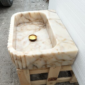 May include: A rectangular marble sink with a fluted edge design. The sink features a cream and brown veined pattern and a gold-colored drain cover. The sink is set on a wooden pallet.