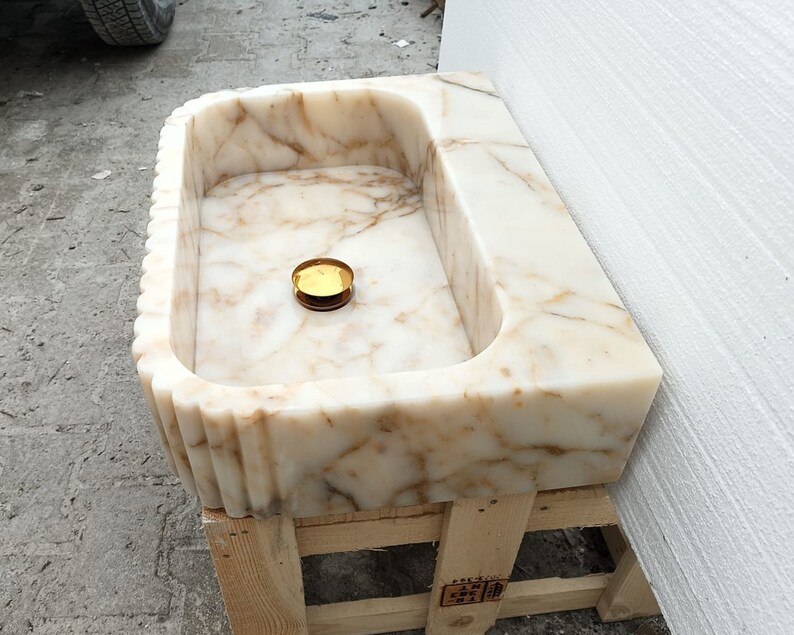 May include: A rectangular marble sink with a fluted edge design. The sink features a cream and brown veined pattern and a gold-colored drain cover. The sink is set on a wooden pallet.