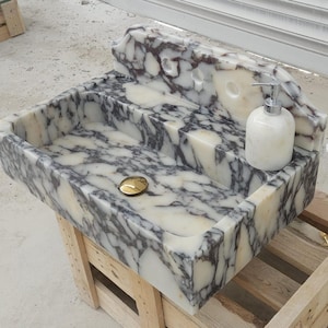 Op de afbeelding: Een rechthoekige marmeren wastafel met een witte basis en donkerpaarse en zwarte aders. De wastafel heeft een verhoogd achterpaneel met drie ronde openingen, een goudkleurige afvoerplug en een witte marmeren zeepdispenser met zilveren pomp.