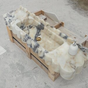 Puede incluir: Un lavabo rectangular de m&aacute;rmol con borde festoneado y vetas blancas y grises. La cubeta incluye un desag&uuml;e dorado y un dispensador de jab&oacute;n blanco sobre la superficie plana. El lavabo se apoya en una estructura de marco de madera.