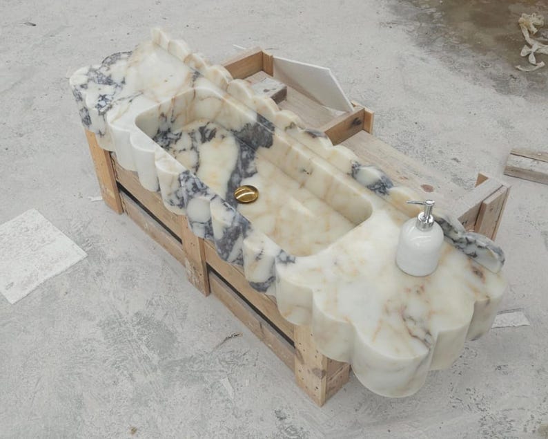 Puede incluir: Un lavabo rectangular de m&aacute;rmol con borde festoneado y vetas blancas y grises. La cubeta incluye un desag&uuml;e dorado y un dispensador de jab&oacute;n blanco sobre la superficie plana. El lavabo se apoya en una estructura de marco de madera.