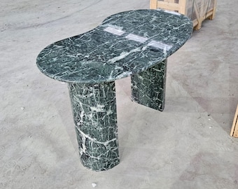 May include: A green marble table with a unique, organic shape. The tabletop is oval, supported by two cylindrical legs and a curved base. The marble has a dark green color with white veining, creating a natural pattern. The table is approximately 40 inches long.
