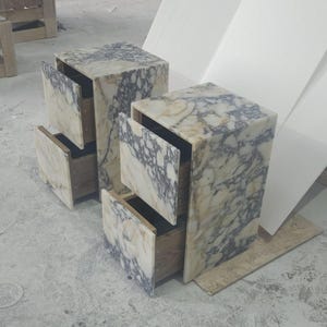 May include: Two marble-patterned nightstands with open drawers. The nightstands are rectangular with a light beige and gray veined pattern. The drawers are open, revealing the interior. The nightstands are on a concrete floor.