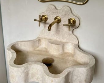 May include: A wall-mounted beige stone sink with a wavy, scalloped edge design. It features a matching stone backsplash and a central tap with two cross-handle controls, all finished in a brushed gold tone. A matching gold drain is visible.