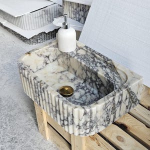 Pode incluir: Uma pia de mármore branco com uma borda canelada, colocada em um suporte de madeira. A pia tem um padrão de veios cinza e uma tampa de drenagem dourada. Um dispensador de sabão branco é colocado na borda da pia.