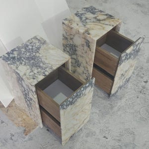 May include: Two marble-patterned bedside tables with open drawers. The tables have a rectangular shape with a light beige and gray color scheme. The drawers are made of wood. The tables are placed on a gray surface.