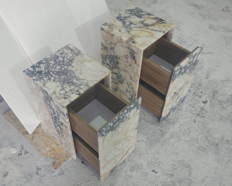 May include: Two marble-patterned bedside tables with open drawers. The tables have a rectangular shape with a light beige and gray color scheme. The drawers are made of wood. The tables are placed on a gray surface.