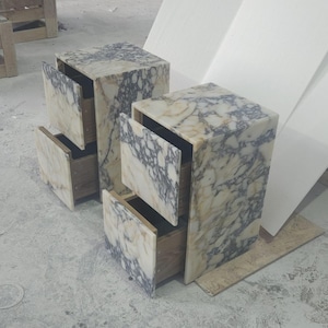 May include: Two marble-patterned nightstands with open drawers. The nightstands are rectangular with a light beige and gray veined pattern. The drawers are open, revealing the wooden interior. The nightstands are on a concrete floor.