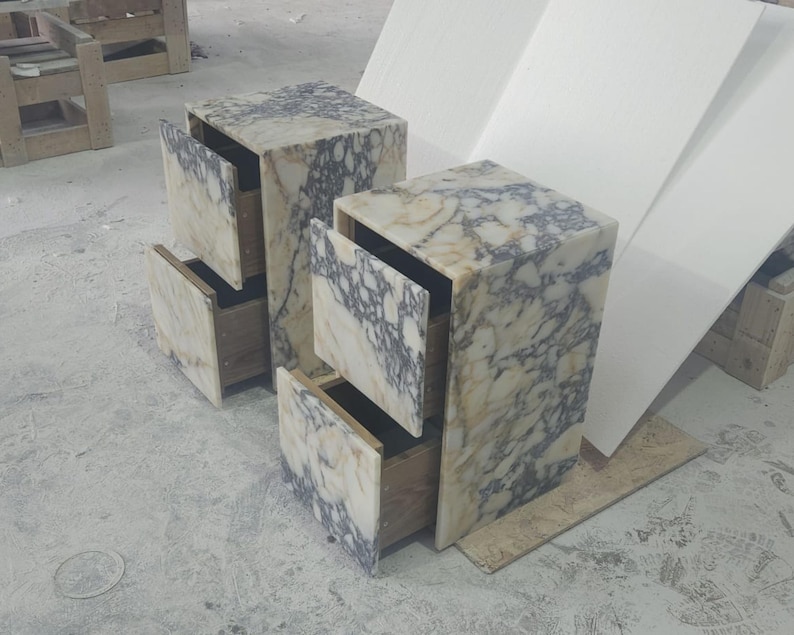 May include: Two marble-patterned nightstands with open drawers. The nightstands are rectangular with a light beige and gray veined pattern. The drawers are open, revealing the wooden interior. The nightstands are on a concrete floor.