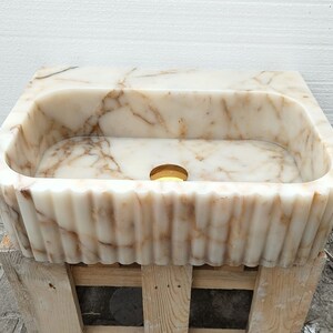 May include: A rectangular, cream-colored marble sink with brown veining. The sink has a fluted, scalloped edge and a circular drain. The sink is set on a wooden pallet.
