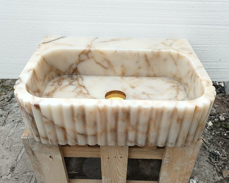 May include: A rectangular, cream-colored marble sink with brown veining. The sink has a fluted, scalloped edge and a circular drain. The sink is set on a wooden pallet.