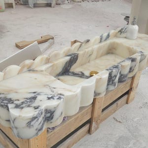 Puede incluir: Un lavabo rectangular de m&aacute;rmol con borde festoneado, base blanca y vetas grises prominentes. El cuenco incluye un desag&uuml;e de tono dorado y un dispensador de jab&oacute;n blanco se encuentra sobre la superficie plana de la encimera de piedra.