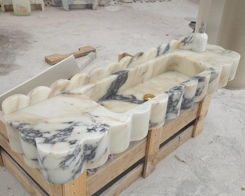 Puede incluir: Un lavabo rectangular de m&aacute;rmol con borde festoneado, base blanca y vetas grises prominentes. El cuenco incluye un desag&uuml;e de tono dorado y un dispensador de jab&oacute;n blanco se encuentra sobre la superficie plana de la encimera de piedra.