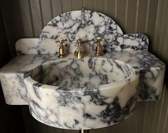 May include: A wall-mounted sink crafted from white marble with prominent dark gray veining. The basin features a rounded front and an integrated backsplash with a curved top. It is equipped with a central brass faucet and two matching brass handles.