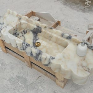 Puede incluir: Un lavabo rectangular de m&aacute;rmol con borde festoneado y vetas blancas y gris oscuro. El cuenco incluye una tapa de desag&uuml;e dorada pulida y un dispensador de jab&oacute;n de cer&aacute;mica blanca sobre la superficie plana de la encimera de piedra.
