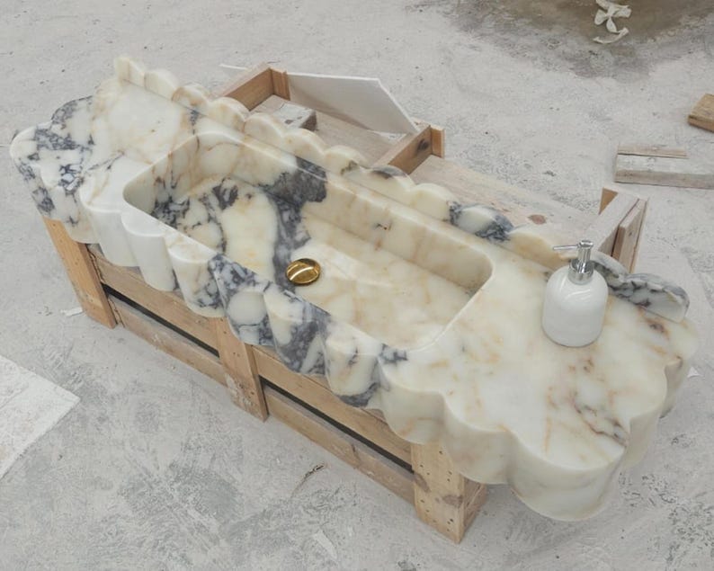 Puede incluir: Un lavabo rectangular de m&aacute;rmol con borde festoneado y vetas blancas y gris oscuro. El cuenco incluye una tapa de desag&uuml;e dorada pulida y un dispensador de jab&oacute;n de cer&aacute;mica blanca sobre la superficie plana de la encimera de piedra.