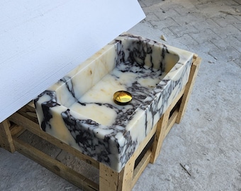 May include: A rectangular stone sink featuring a polished cream and white base with prominent dark purple and black veining throughout the basin and exterior walls. A circular, reflective gold-toned metal drain cover is centered at the bottom.