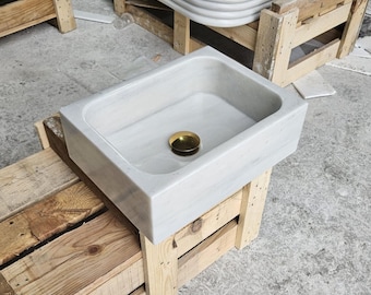 May include: A rectangular white marble sink featuring a smooth, polished finish and a centred circular brass drain cover. The basin has thick, solid walls and a deep, open interior, showcasing the natural light grey veining of the stone material.
