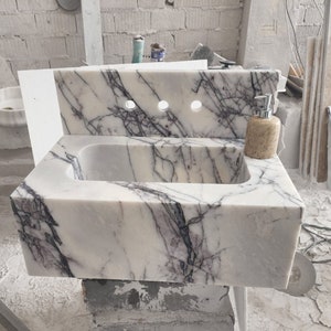 Puede incluir: Un lavabo de baño de mármol blanco con vetas moradas. El lavabo está montado en una pared y tiene una forma rectangular. Hay tres agujeros en el salpicadero para los accesorios.