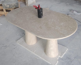 May include: An oval-shaped, beige stone table with two cylindrical pedestal legs. A small black vase with red floral accents sits on the table's surface. The table is a neutral color and appears to be made of stone.