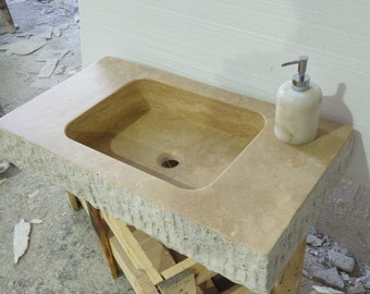 May include: A rectangular beige stone sink with a smooth basin and a rough, chiseled front edge. The sink features a central drain hole and is topped with a white, cylindrical soap dispenser with a silver pump mechanism, resting on a wooden frame.