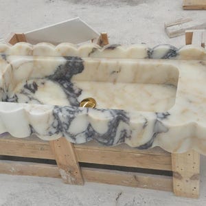 Puede incluir: Un lavabo rectangular de m&aacute;rmol con borde festoneado y vetas blancas y gris oscuro. La cubeta incluye un desag&uuml;e dorado y un dispensador de jab&oacute;n blanco. El lavabo se apoya sobre una base de madera utilizada para su transporte.