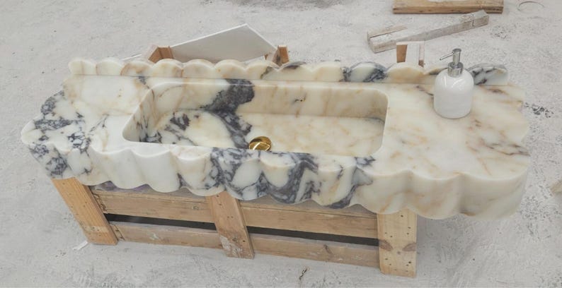 Puede incluir: Un lavabo rectangular de m&aacute;rmol con borde festoneado y vetas blancas y gris oscuro. La cubeta incluye un desag&uuml;e dorado y un dispensador de jab&oacute;n blanco. El lavabo se apoya sobre una base de madera utilizada para su transporte.