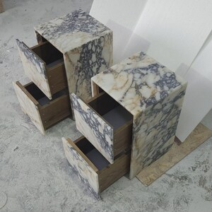 May include: Two marble-topped nightstands with open drawers. The marble has a cream and gray veined pattern. The drawers are made of wood. The nightstands are rectangular and sit on a concrete floor.