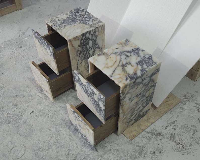 May include: Two marble-topped nightstands with open drawers. The marble has a cream and gray veined pattern. The drawers are made of wood. The nightstands are rectangular and sit on a concrete floor.