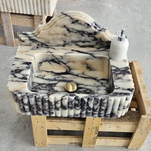 Op de afbeelding: Een rechthoekige marmeren wastafel met een crème en zwart geaderd patroon, een geschulpte voorrand en een hoge achterwand. De wasbak is voorzien van een centrale goudkleurige afvoerplug en een witte zeepdispenser in de rechterbovenhoek.