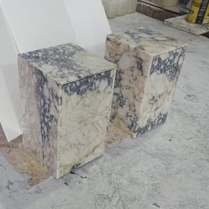 May include: Two rectangular marble blocks with a cream and gray veined pattern. The blocks are upright and have a box-like shape, possibly intended as pedestals or small tables. The marble has a polished finish.