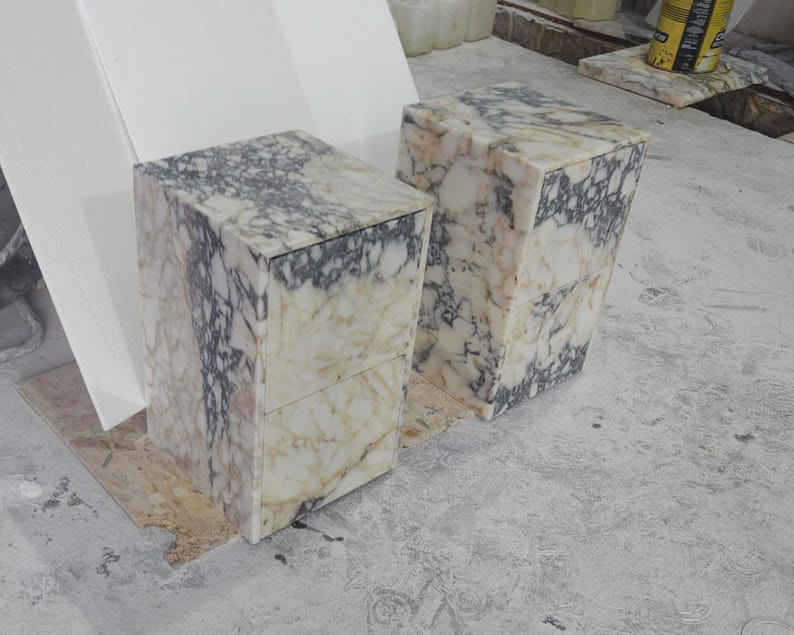 May include: Two rectangular marble blocks with a cream and gray veined pattern. The blocks are upright and have a box-like shape, possibly intended as pedestals or small tables. The marble has a polished finish.