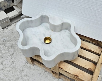 May include: A white marble sink with a wavy, organic shape and a polished gold-toned drain cover. The basin features natural grey veining throughout the stone surface, creating a distinct pattern. The sink has a smooth, rounded rim and deep interior.