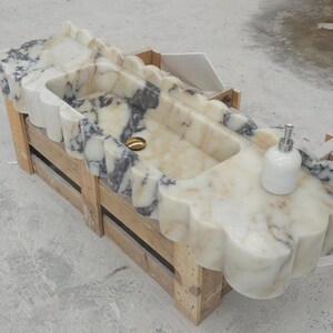 Puede incluir: Un lavabo rectangular de m&aacute;rmol con borde festoneado y vetas en crema, blanco y gris oscuro. La cubeta incluye una tapa de desag&uuml;e dorada y un dispensador de jab&oacute;n blanco sobre la superficie, apoyado sobre una caja de madera de transporte.