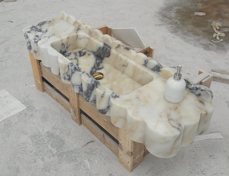 Puede incluir: Un lavabo rectangular de m&aacute;rmol con borde festoneado y vetas en crema, blanco y gris oscuro. La cubeta incluye una tapa de desag&uuml;e dorada y un dispensador de jab&oacute;n blanco sobre la superficie, apoyado sobre una caja de madera de transporte.